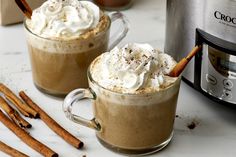 two mugs of hot chocolate with whipped cream and cinnamon sticks next to an air fryer