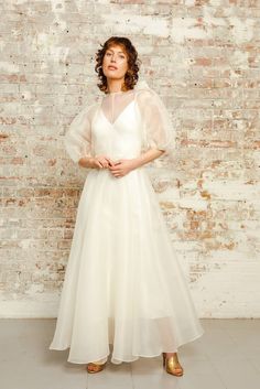 a woman standing in front of a brick wall wearing a white dress and gold shoes