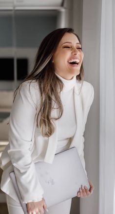 a woman laughing and holding a laptop computer in her right hand while standing next to a window