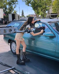 a woman in short skirt and boots standing next to a blue car holding a camera