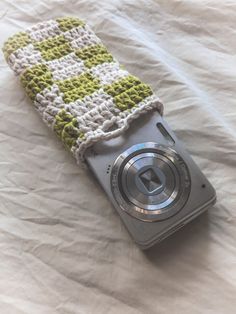 a camera sitting on top of a bed next to a white sheet with a green and white crocheted cover