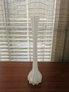 a white vase sitting on top of a wooden table next to a window covered in blinds