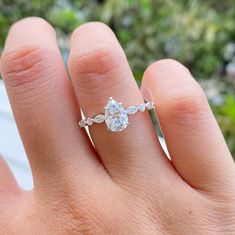 a woman's hand with a diamond ring on top of her finger, in front of some trees