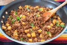 a skillet filled with meat and potatoes on top of a colorful table cloth next to a wooden spoon