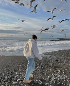 a woman standing on top of a rocky beach next to the ocean with seagulls flying overhead