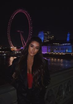 a woman standing in front of a ferris wheel at night with the city lights behind her