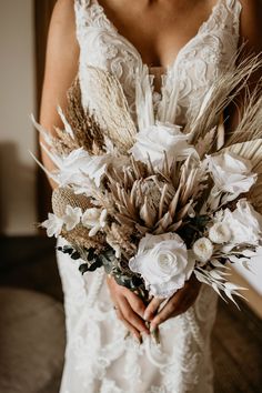 a woman in a wedding dress holding a bouquet of dried flowers and feathers on her arm