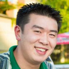 a man with black hair and green shirt smiling at the camera in front of a building