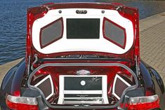 the back end of a red and white sports car with its trunk open in front of a body of water