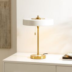 a table lamp sitting on top of a white dresser next to a book and phone