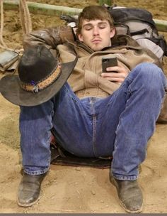 a man sitting on the ground with his cell phone in his hand and wearing a cowboy hat