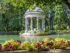 a gazebo in the middle of a pond surrounded by trees and shrubs with flowers around it