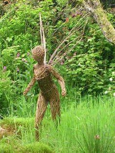 a statue made out of branches in the middle of some grass and bushes with trees behind it