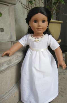 the doll is wearing a white dress and gold earrings, sitting on a ledge next to a potted plant