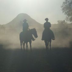 two people riding horses in front of a mountain with mist coming off the ground behind them