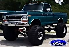 a large green truck with big tires parked in a parking lot next to some trees