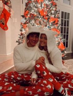 two people sitting in front of a christmas tree wearing matching pajamas and hoodies with santa claus on them