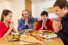 a group of people sitting around a table with food on it and praying in front of them