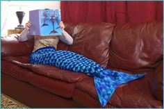 a woman sitting on a couch while reading a book with a mermaid tail blanket over her head