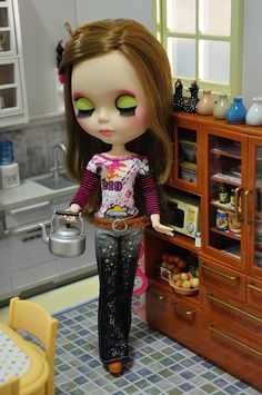 a doll with green eyes is holding a teapot in her hand and standing next to a wooden cabinet