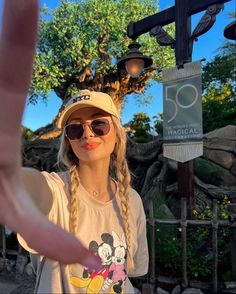 a woman in sunglasses and a hat making a peace sign with her hand while standing next to a tree