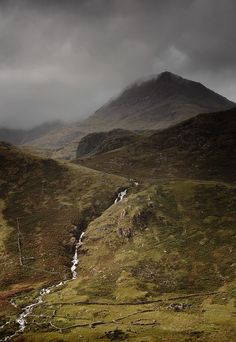 a small stream running through the middle of a lush green mountain valley under a dark cloudy sky