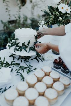 two people are decorating a white cake and cupcakes on a table with greenery