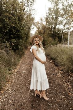 a pregnant woman in a white dress and hat standing on a dirt road