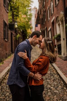 a man and woman kissing in an alleyway with brick buildings on either side as well as cobblestones