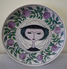 a decorative plate with a woman's face painted on the front and purple roses around it