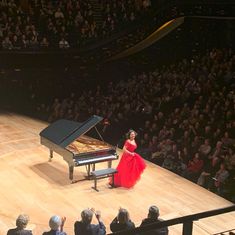 a woman in a red dress standing next to a piano on a stage with an audience