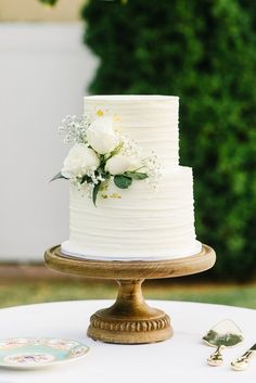 a wedding cake with white flowers and greenery sits on a wooden stand in front of a green bush