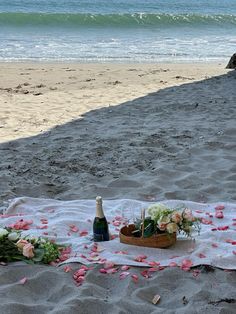 a bottle of champagne and flowers on a blanket at the beach with petals in the sand