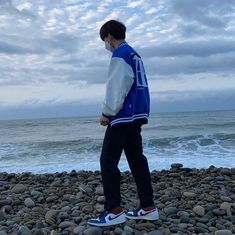 a man standing on top of a pebble covered beach next to the ocean wearing a blue and white jacket