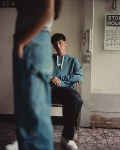 a young man sitting on a chair in front of a door with his legs crossed