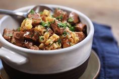 a white bowl filled with pasta and meat on top of a blue cloth next to a spoon