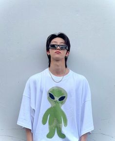 a young man wearing sunglasses and an alien t - shirt stands against a white wall