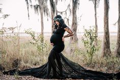 a pregnant woman wearing a black gown and headpiece stands in the woods with her hands on her hips