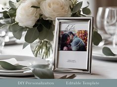 a table setting with white flowers and greenery in the center, including an empty photo frame