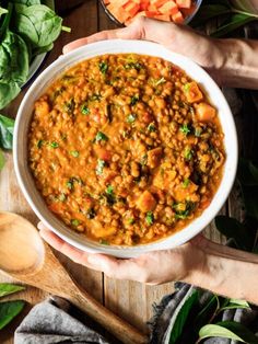 two hands holding a bowl of lentils and carrots with spinach leaves on the side