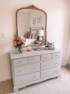 a white dresser topped with a mirror next to a bed and vase filled with flowers