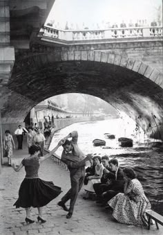 an old black and white photo of people dancing in front of a river under a bridge