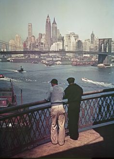 two men standing on a bridge looking out at the water and buildings in the background