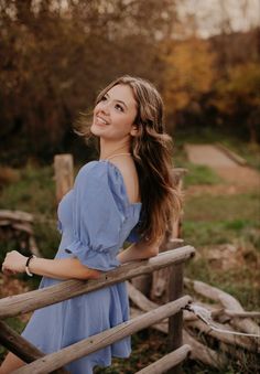 a woman in a blue dress leaning on a wooden fence with her hair blowing in the wind