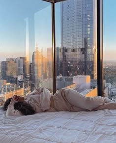 a woman laying on top of a bed in front of a large window with city view