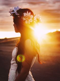 a woman with flowers in her hair is standing on the beach at sundown, looking off into the distance