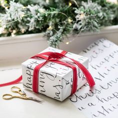 a present wrapped in white paper with red ribbon sitting on top of a table next to a christmas tree