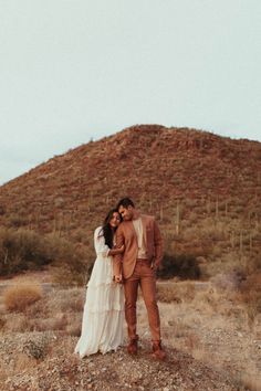 a bride and groom standing in front of a mountain with their arms around each other