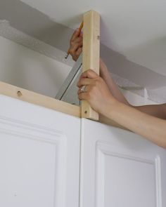 a person holding a piece of wood up to the top of a cabinet with a pair of scissors