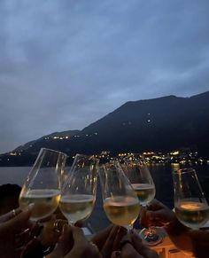 several people toasting with wine glasses in front of a lake at night, while the sun is setting